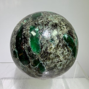 May include: A spherical gemstone with a polished surface, displaying a mix of emerald green, black, and grey hues. The stone's surface is smooth and rounded, with a glossy finish that highlights its natural patterns.