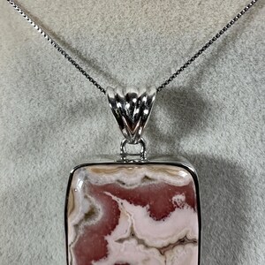 May include: A rectangular pendant necklace with a silver chain. The pendant features a polished stone with a pattern of pink and cream swirls, set in a silver frame. The pendant is approximately 1.5 inches long.