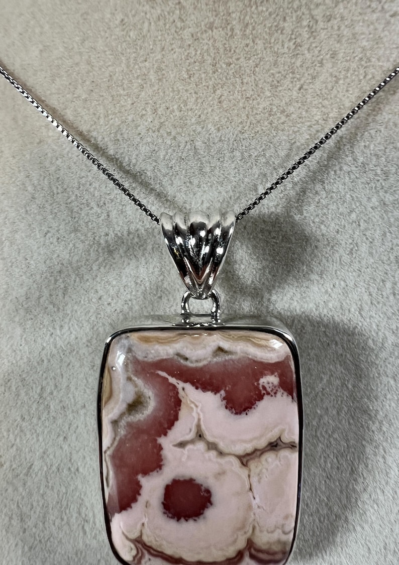 May include: A rectangular pendant necklace with a silver chain. The pendant features a polished stone with a pattern of pink and cream swirls, set in a silver frame. The pendant is approximately 1.5 inches long.