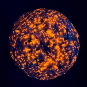 Puede incluir: Un espécimen de roca esférico que muestra intensas manchas fluorescentes de color naranja y amarillo sobre una superficie oscura y texturizada. El mineral presenta un aspecto moteado e irregular con grietas y protuberancias.
