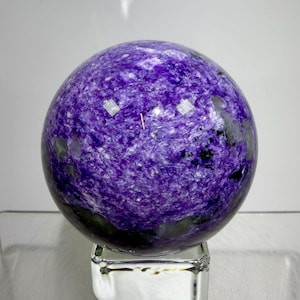 May include: A polished spherical charoite gemstone featuring a vibrant purple colour with swirling white and black mineral inclusions. The stone is displayed resting on a clear, square glass stand, showcasing a smooth, reflective surface.