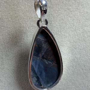 May include: A teardrop-shaped pendant with a polished silver setting and chain. The gemstone within the setting displays a mix of dark blue, black, and reddish-brown hues. The pendant is suspended from a delicate silver chain, perfect for everyday wear.