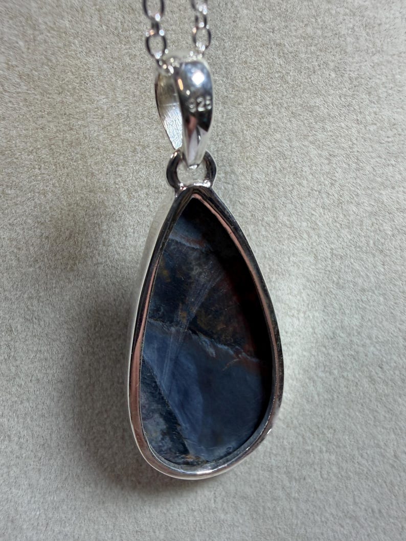 May include: A teardrop-shaped pendant with a polished silver setting and chain. The gemstone within the setting displays a mix of dark blue, black, and reddish-brown hues. The pendant is suspended from a delicate silver chain, perfect for everyday wear.