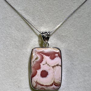 May include: A rectangular pendant necklace featuring a polished stone with a pink and white pattern. The stone is set in a silver-colored metal frame and hangs from a delicate silver chain. The pendant has a unique, organic design.