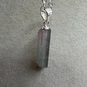 May include: A silver chain necklace featuring a rectangular, multi-coloured crystal pendant. The crystal displays green, grey, and purple tones. The pendant is connected to the chain with a silver bail. The background is a plain grey colour.