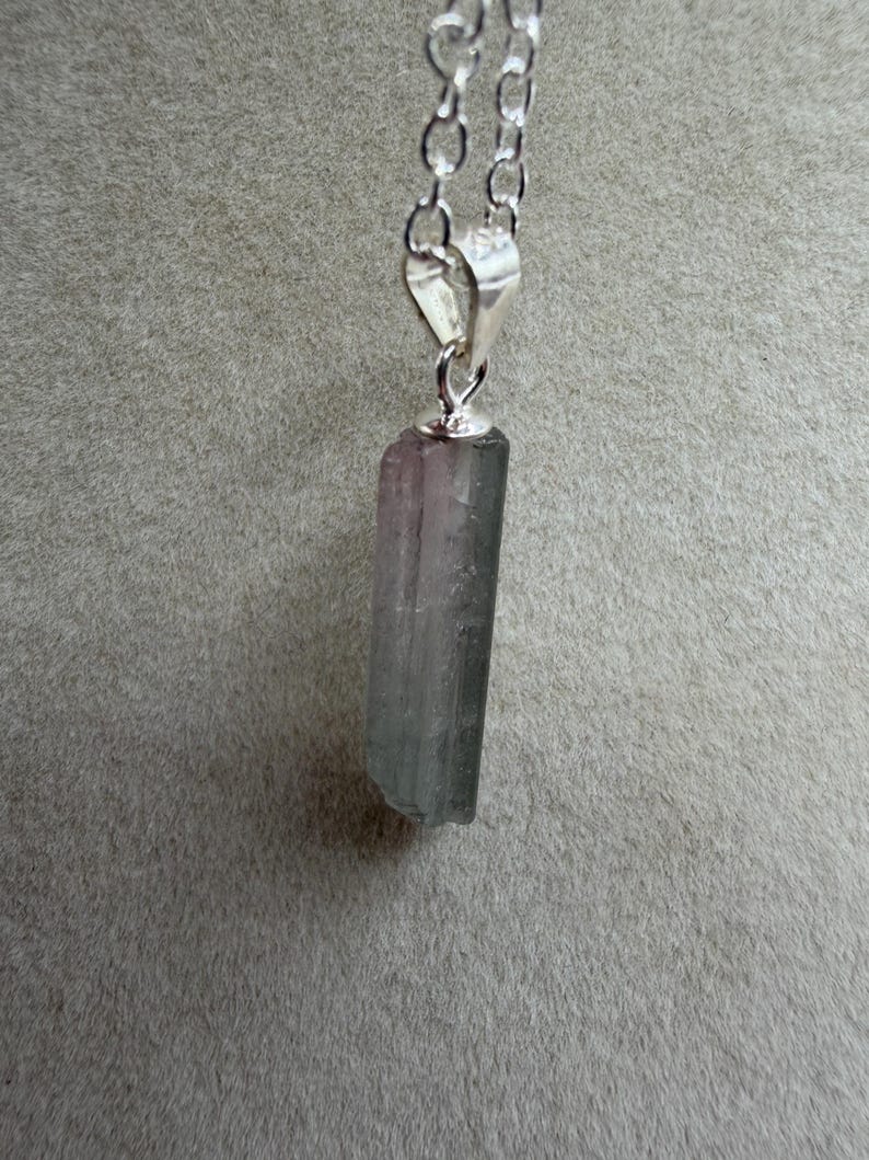 May include: A silver chain necklace featuring a rectangular, multi-coloured crystal pendant. The crystal displays green, grey, and purple tones. The pendant is connected to the chain with a silver bail. The background is a plain grey colour.