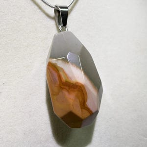 May include: A gemstone pendant necklace with a silver chain. The pendant is a faceted, irregular shape with a mix of colors, including gray, brown, and pink. The necklace is displayed against a white background.