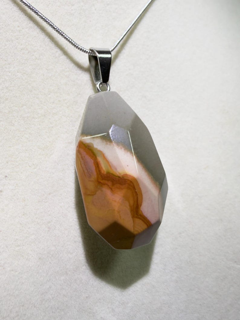 May include: A gemstone pendant necklace with a silver chain. The pendant is a faceted, irregular shape with a mix of colors, including gray, brown, and pink. The necklace is displayed against a white background.