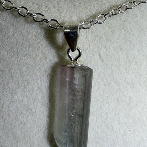 May include: A pendant necklace featuring a raw, elongated, translucent crystal with a gradient of pale pink at the top transitioning into a dark grey base. The crystal is held by a silver-toned metal bail attached to a matching silver-toned chain.