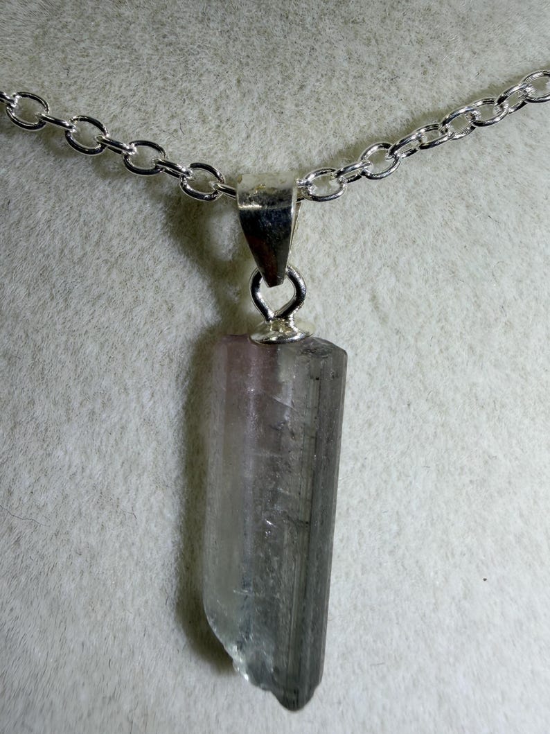 May include: A pendant necklace featuring a raw, elongated, translucent crystal with a gradient of pale pink at the top transitioning into a dark grey base. The crystal is held by a silver-toned metal bail attached to a matching silver-toned chain.