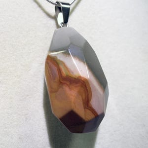 May include: A faceted pendant necklace featuring a large, irregularly shaped stone. The stone displays a mix of colors, including gray, brown, and burgundy, with a polished surface. The pendant hangs from a silver chain, creating a striking contrast.