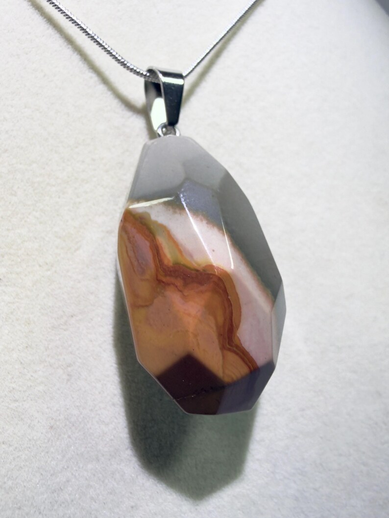 May include: A faceted pendant necklace featuring a large, irregularly shaped stone. The stone displays a mix of colors, including gray, brown, and burgundy, with a polished surface. The pendant hangs from a silver chain, creating a striking contrast.