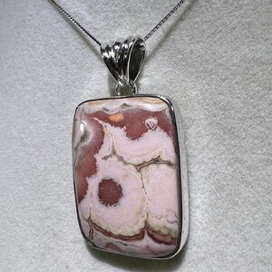 May include: A rectangular pendant with a silver-colored frame and bail, suspended from a delicate chain. The pendant features a polished stone with a swirling pattern of pink, white, and brown hues, creating a unique, natural design. The necklace is a piece of jewelry.