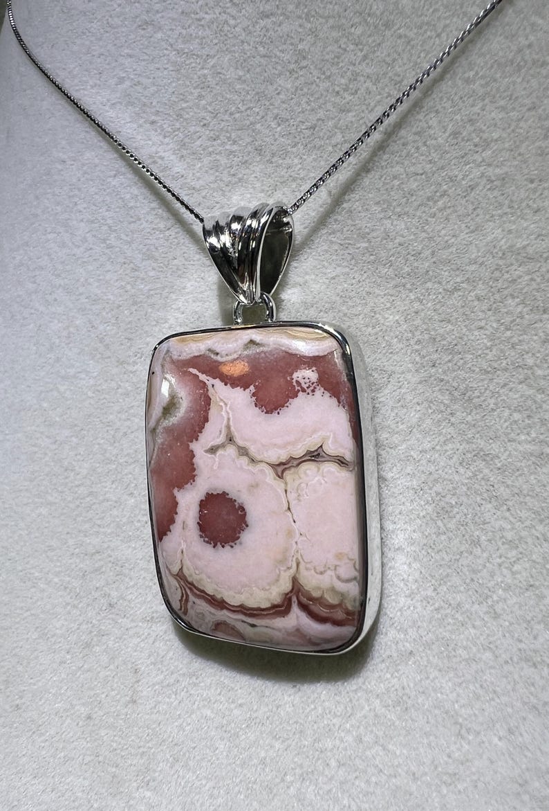 May include: A rectangular pendant with a silver-colored frame and bail, suspended from a delicate chain. The pendant features a polished stone with a swirling pattern of pink, white, and brown hues, creating a unique, natural design. The necklace is a piece of jewelry.