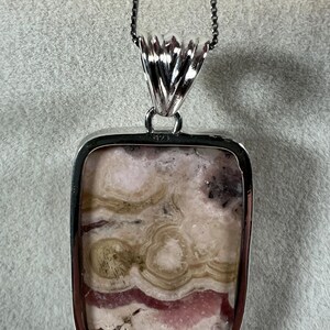 May include: A silver pendant necklace with a rectangular stone. The stone has a mix of pink, beige, and brown colors with a unique pattern. The pendant is attached to a silver chain, creating a stylish accessory.