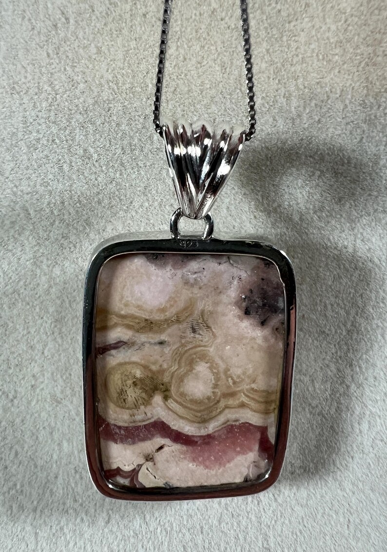May include: A silver pendant necklace with a rectangular stone. The stone has a mix of pink, beige, and brown colors with a unique pattern. The pendant is attached to a silver chain, creating a stylish accessory.