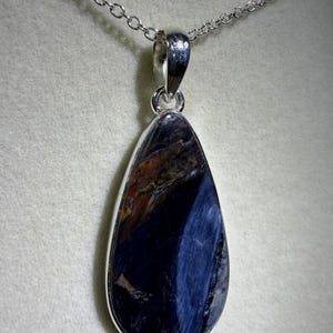 May include: A teardrop-shaped pendant with a polished, dark blue gemstone, accented with streaks of lighter blue and brown. The stone is set in a silver-colored metal frame and hangs from a delicate silver chain. The necklace is displayed against a neutral background.