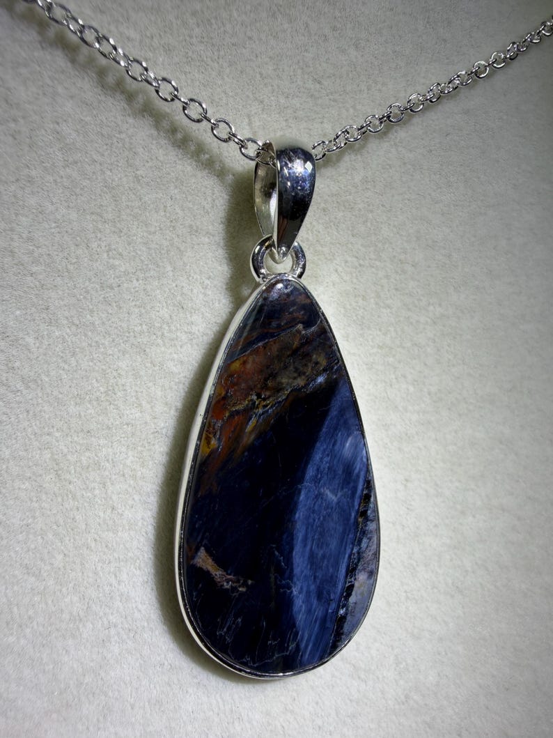 May include: A teardrop-shaped pendant with a polished, dark blue gemstone, accented with streaks of lighter blue and brown. The stone is set in a silver-colored metal frame and hangs from a delicate silver chain. The necklace is displayed against a neutral background.