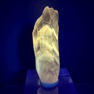 May include: A translucent, pale yellow crystalline mineral specimen with a jagged, elongated vertical structure. The crystal features natural facets and internal fractures, illuminated to glow against a dark, deep blue background.