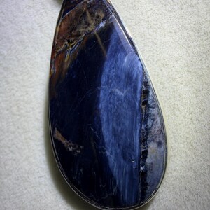 May include: A teardrop-shaped pendant featuring a polished gemstone with a silver-colored metal border. The stone displays a mix of deep blue, brown, and white colors, creating a unique pattern. The pendant is suspended from a silver-colored chain.