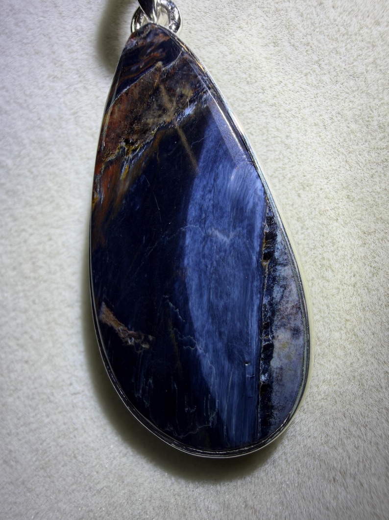 May include: A teardrop-shaped pendant featuring a polished gemstone with a silver-colored metal border. The stone displays a mix of deep blue, brown, and white colors, creating a unique pattern. The pendant is suspended from a silver-colored chain.