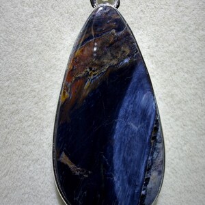 May include: A teardrop-shaped pendant with a polished, dark blue gemstone. The stone has streaks of brown, orange, and lighter blue, set in a silver-colored metal frame. The pendant is designed to be worn as a necklace.