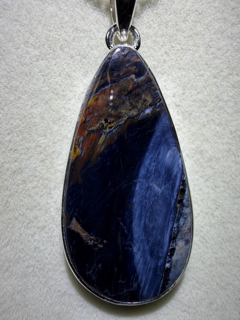 May include: A teardrop-shaped pendant with a polished, dark blue gemstone. The stone has streaks of brown, orange, and lighter blue, set in a silver-colored metal frame. The pendant is designed to be worn as a necklace.