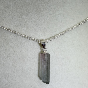 May include: A silver necklace with a pendant made from a grey and pink tourmaline crystal. The crystal is rectangular with a natural, uneven edge. The chain is a delicate silver link.