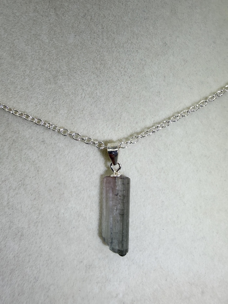 May include: A silver necklace with a pendant made from a grey and pink tourmaline crystal. The crystal is rectangular with a natural, uneven edge. The chain is a delicate silver link.