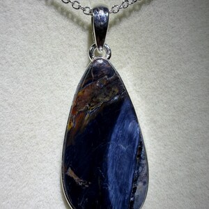 May include: A teardrop-shaped pendant necklace featuring a polished gemstone with a silver-colored metal setting. The gemstone displays a mix of dark blue, brown, and orange hues, with a silver chain.