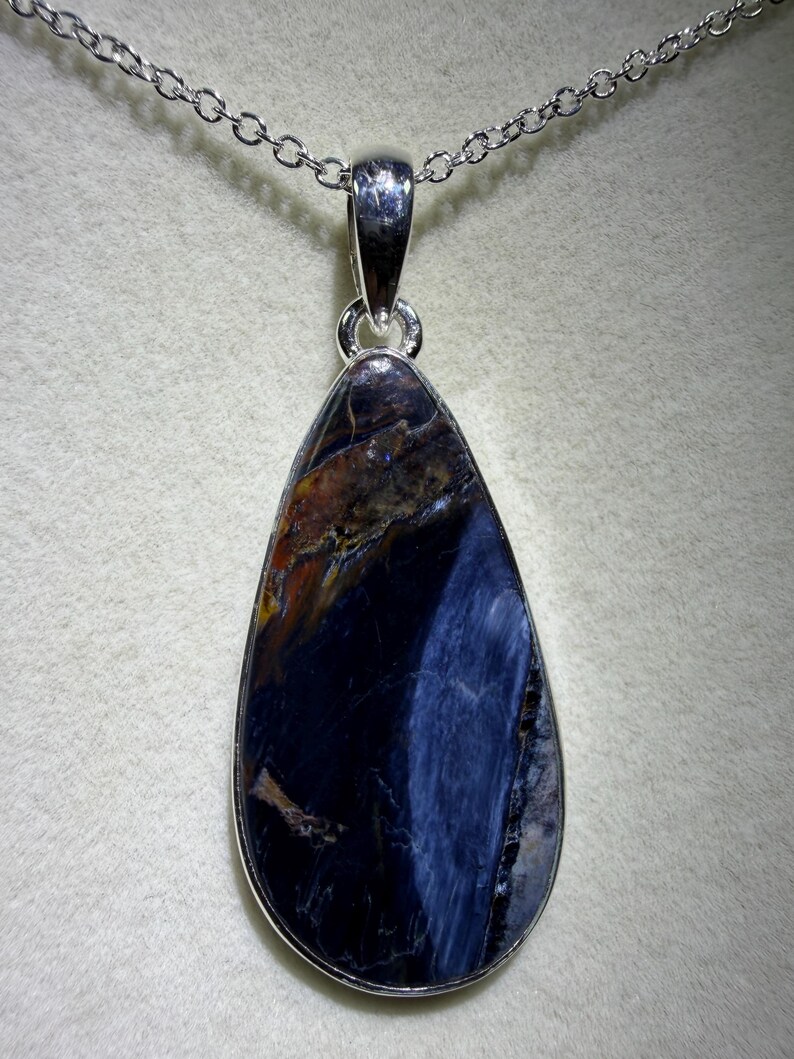 May include: A teardrop-shaped pendant necklace featuring a polished gemstone with a silver-colored metal setting. The gemstone displays a mix of dark blue, brown, and orange hues, with a silver chain.