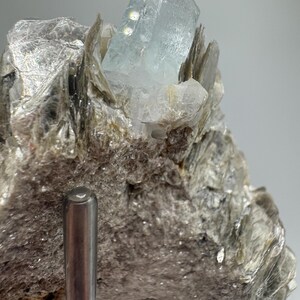 May include: A light blue aquamarine crystal on a bed of gray mica. The crystal is clear and has a slight green tint. The mica is rough and textured.