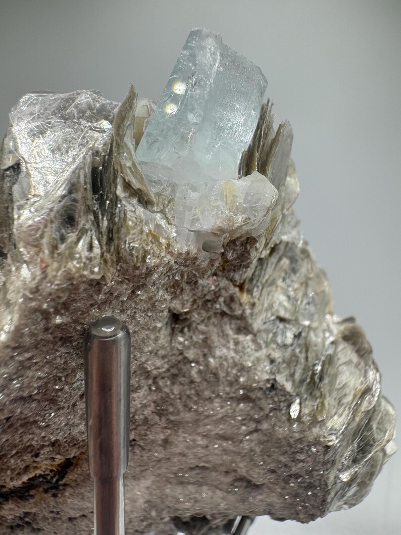 May include: A light blue aquamarine crystal on a bed of gray mica. The crystal is clear and has a slight green tint. The mica is rough and textured.