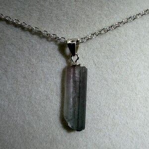 May include: A silver chain necklace featuring a natural tourmaline pendant. The pendant is a vertical, rectangular crystal with pink, grey, and green hues. The chain is a delicate link design.