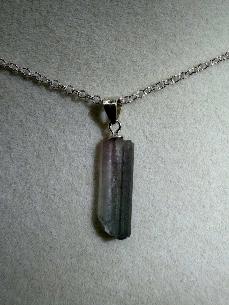 May include: A silver chain necklace featuring a natural tourmaline pendant. The pendant is a vertical, rectangular crystal with pink, grey, and green hues. The chain is a delicate link design.