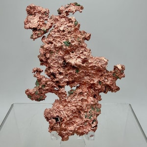 May include: A raw, metallic copper specimen featuring a jagged, irregular, and porous structure with a distinct reddish-brown lustre. The surface displays small, scattered patches of green mineral deposits embedded within the textured, crystalline form.