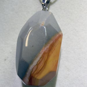 May include: A faceted pendant necklace featuring a large, polished stone with a mix of colors. The stone has a gray, blue, brown, and yellow color scheme. The pendant is suspended from a silver-colored chain.