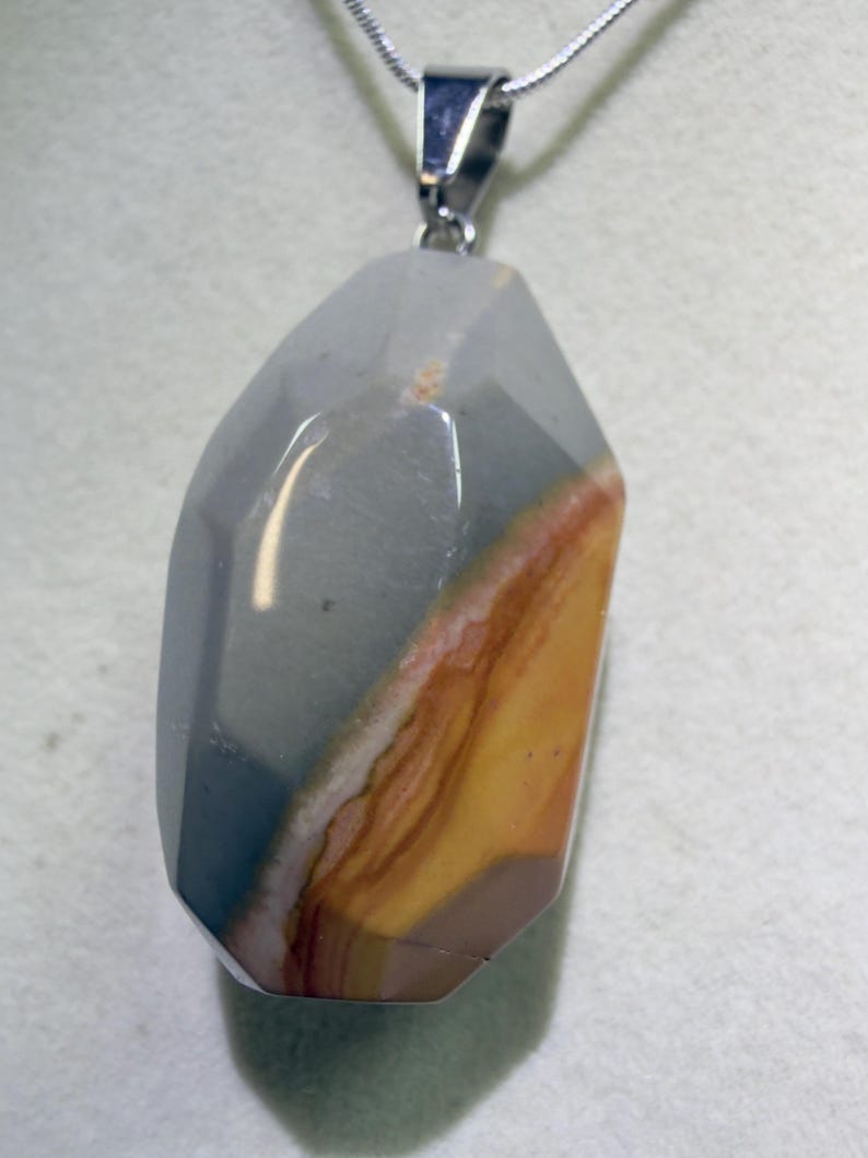 May include: A faceted pendant necklace featuring a large, polished stone with a mix of colors. The stone has a gray, blue, brown, and yellow color scheme. The pendant is suspended from a silver-colored chain.