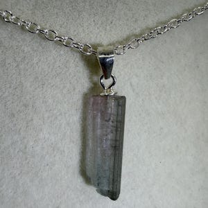 May include: A silver chain necklace featuring a rectangular tourmaline pendant. The pendant displays a blend of pink, grey, and clear hues. The tourmaline measures approximately 2.5 cm in length and is suspended from a fine silver chain.