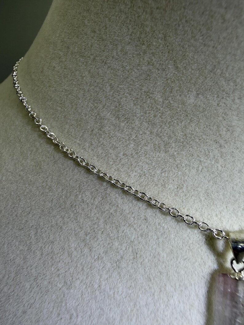 May include: A silver-toned metal chain necklace featuring a delicate link design. The chain is attached to a metallic bail that holds a vertical, translucent, pale pink rectangular gemstone pendant with a smooth, polished surface.