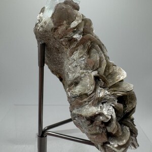 May include: A light blue aquamarine crystal perched atop a cluster of silvery-grey mica crystals. The specimen is mounted on a black metal stand.