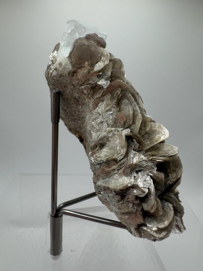 May include: A light blue aquamarine crystal perched atop a cluster of silvery-grey mica crystals. The specimen is mounted on a black metal stand.