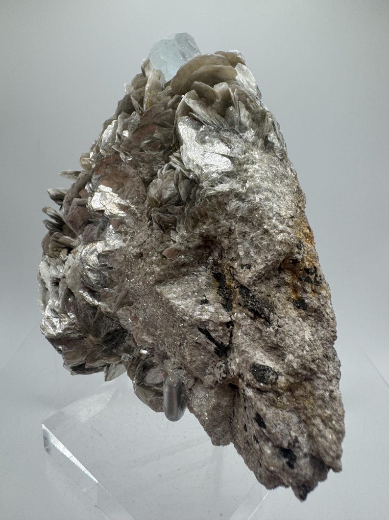May include: A cluster of silvery-grey mica crystals with a light blue aquamarine crystal on top. The crystals are on a white background.