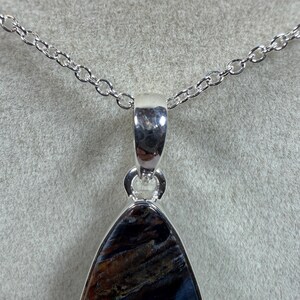 May include: A silver pendant necklace featuring a teardrop-shaped gemstone with a mix of dark blue, brown, and gold tones. The pendant is suspended from a delicate silver chain. The gemstone has a polished finish.