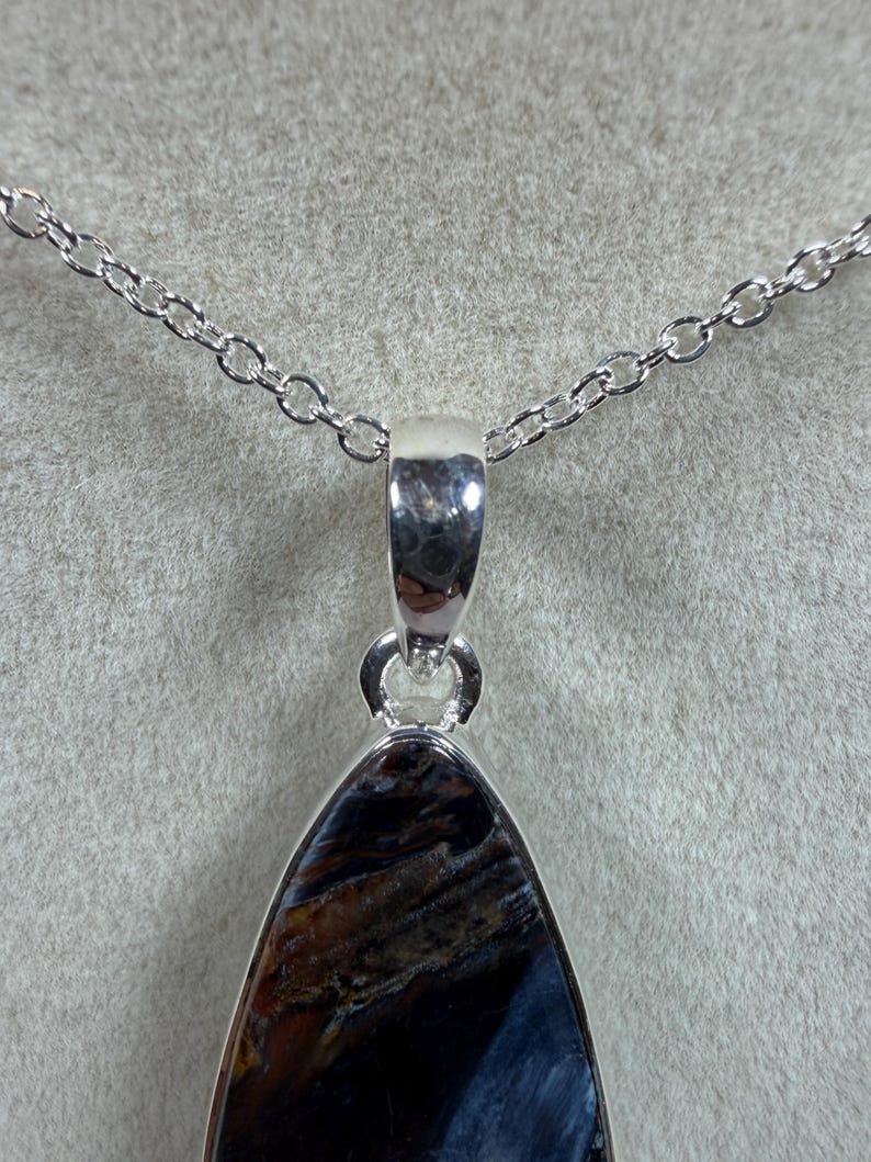 May include: A silver pendant necklace featuring a teardrop-shaped gemstone with a mix of dark blue, brown, and gold tones. The pendant is suspended from a delicate silver chain. The gemstone has a polished finish.