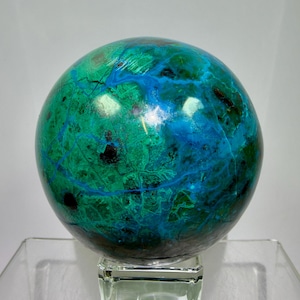 Puede incluir: Una piedra preciosa esférica y pulida que presenta una mezcla vibrante de verde bosque profundo, turquesa brillante y vetas azul eléctrico. La piedra tiene una superficie lisa y brillante con inclusiones oscuras sobre una base de cristal.