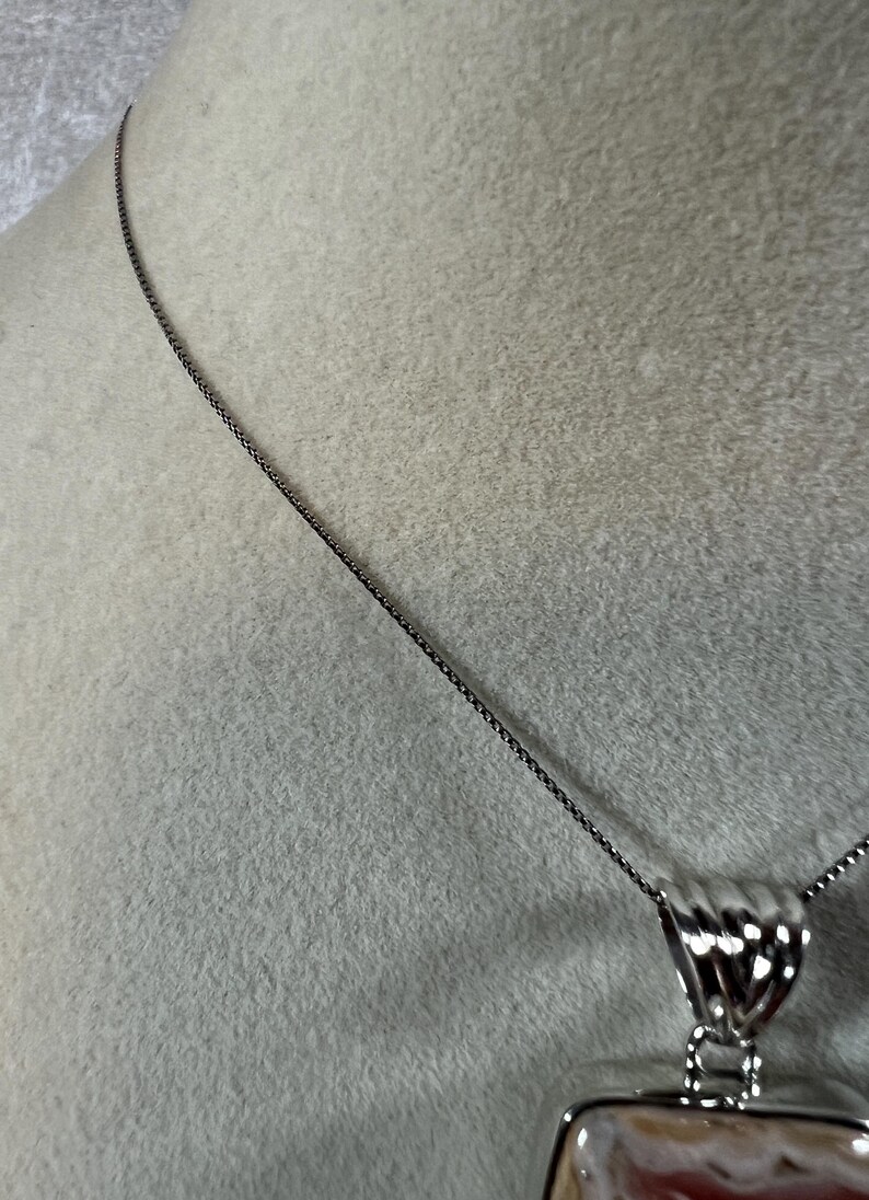 May include: A silver necklace with a rectangular pendant. The pendant features a mix of colors, including pink, white, and brown. The necklace chain is a dark silver color, and the pendant has a decorative silver bail.