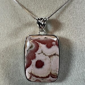 May include: A silver necklace featuring a rectangular pendant with a unique pattern of pink and white swirls. The pendant is encased in a silver frame and hangs from a delicate silver chain.