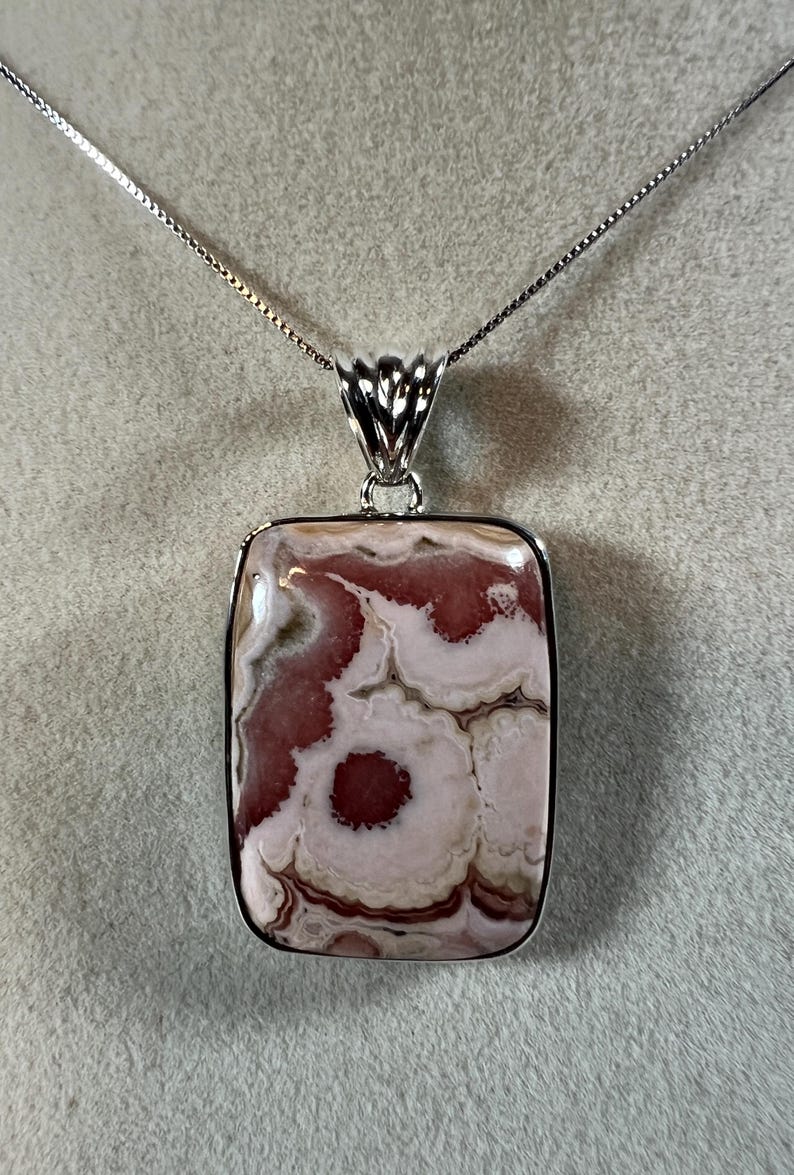 May include: A silver necklace featuring a rectangular pendant with a unique pattern of pink and white swirls. The pendant is encased in a silver frame and hangs from a delicate silver chain.