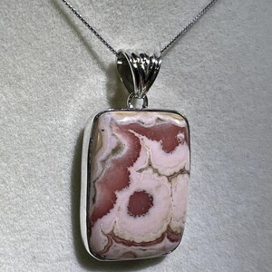 May include: A rectangular pendant with a silver-colored bezel and chain. The pendant features a polished stone with a pattern of pink and white swirls. The stone has a unique, natural appearance, and the chain is delicate.